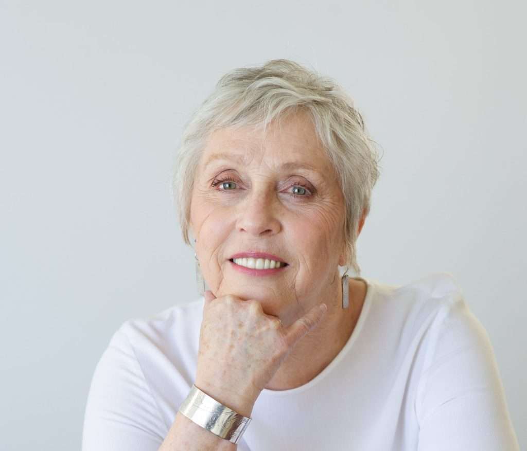 Hélène Grandmaître, member of the Embrun Historical Society committee, smiling warmly while resting her chin on her hand. She is wearing a white top, silver bracelet, and earrings, seated against a light background.